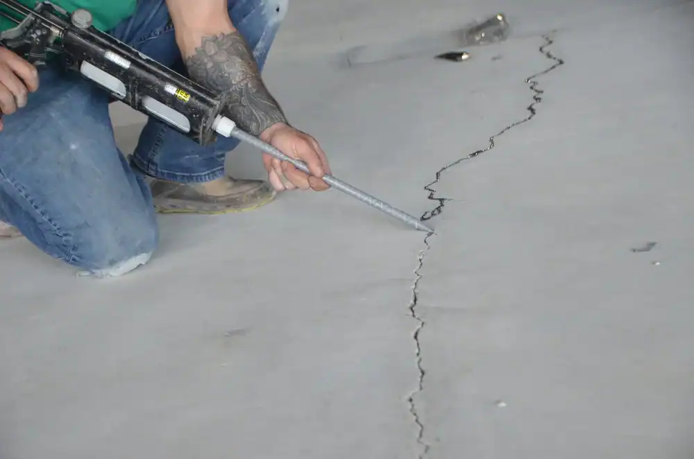 A person kneels on a concrete floor in Middlesex & New London County, CT, using a caulking gun to fill a long, jagged crack&mdash;prepping the surface for epoxy flooring. Their tattooed arm, blue jeans, and work boots show they're ready for the job.