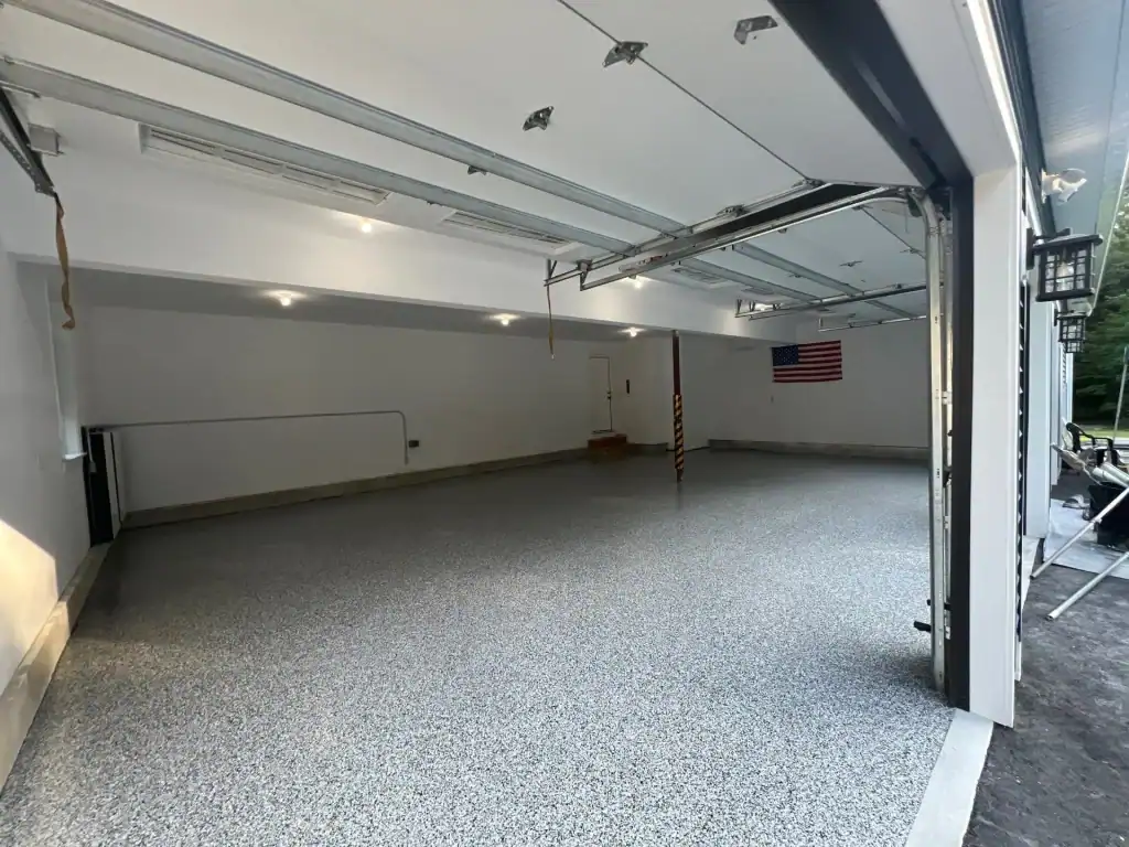 A clean, spacious garage with epoxy flooring in Hartford, CT, features white walls, overhead lighting, and an American flag on the far wall. The open doors let in daylight&mdash;perfect for homes in Middlesex & New London County.