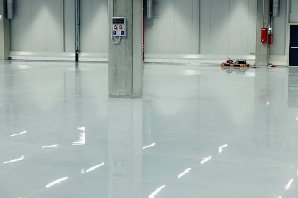 Large, empty industrial warehouse with smooth, glossy, light gray epoxy flooring in Middlesex & New London County, CT; a concrete pillar and wall-mounted control panel are visible, with some equipment and pipes along the walls in the background.