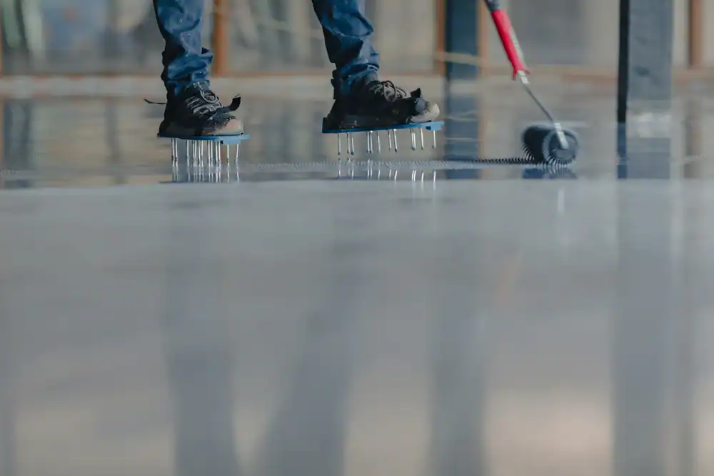 A person wearing spiked shoes uses a roller to spread epoxy on a shiny floor inside a building, perfect for epoxy flooring projects in Hartford, Middlesex & New London County. Only the legs and feet are visible.