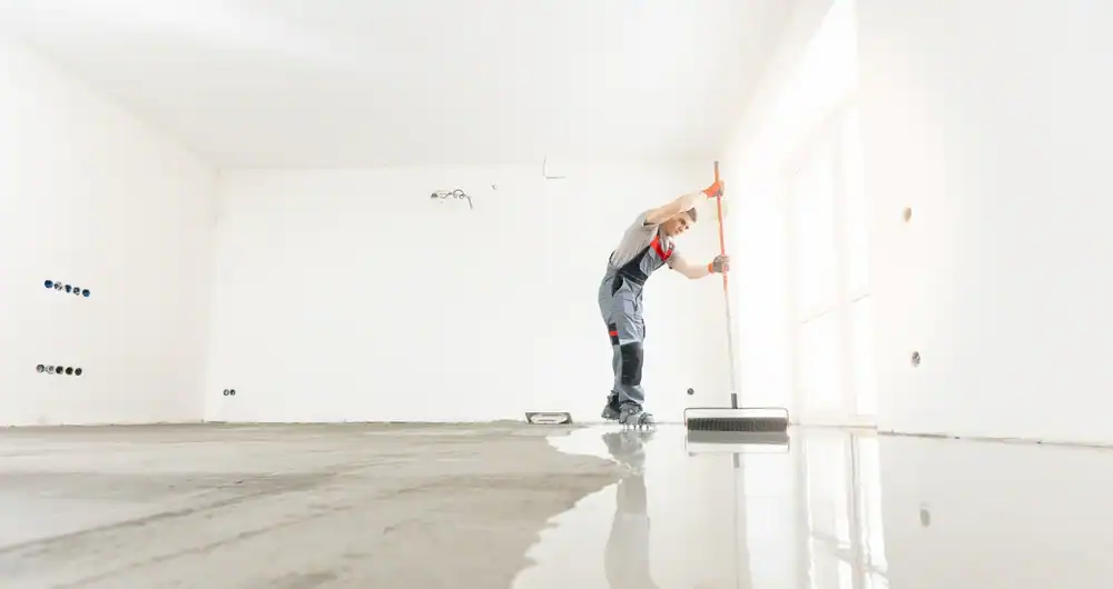 A worker in overalls uses a long-handled roller to coat the floor of an empty, bright, white room, possibly applying an epoxy flooring finish often seen in Hartford, CT and New Haven.