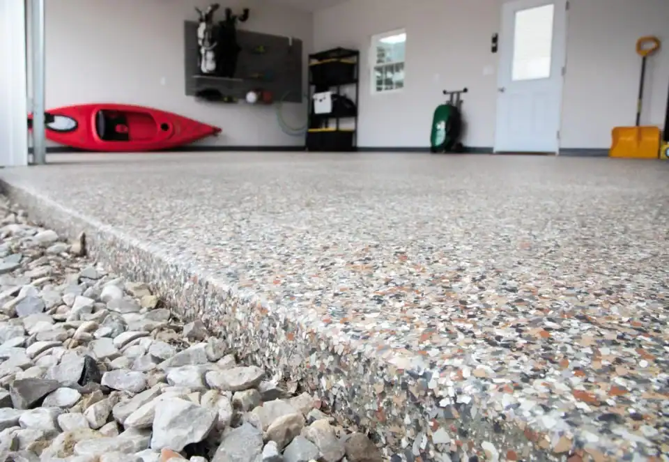 Close-up of a polished epoxy flooring garage in Hartford, CT, with an exposed aggregate edge leading to a gravel area. Inside, a red kayak, tools, and shelves line the back wall&mdash;showcasing stylish storage in Middlesex & New London County.