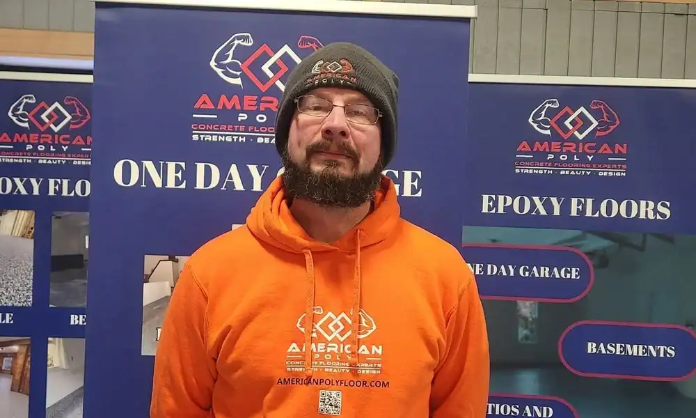 A man with a beard and glasses wears an orange "American Poly" hoodie and gray beanie, standing in front of banners advertising professional epoxy flooring East Hartford, CT, and concrete services.