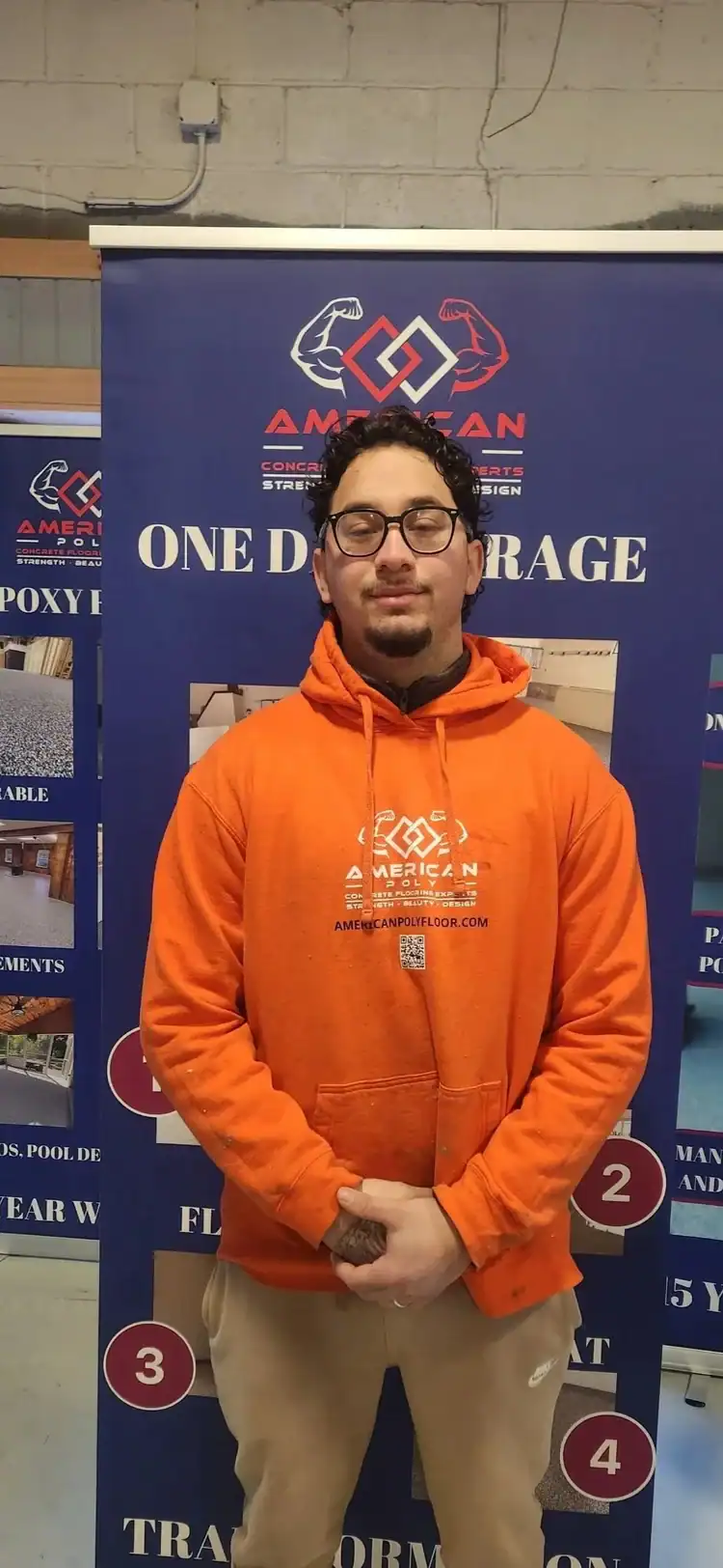 A man wearing glasses and an orange hoodie stands in front of a blue promotional banner for American Garage Floor Coatings, featuring text and graphics about epoxy flooring Hartford, CT. He stands with his hands clasped and smiles slightly.