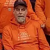 A man with a gray beard wearing an orange hoodie and a dark cap is sitting and smiling in New Haven, CT. Other people in matching orange hoodies, possibly part of an epoxy flooring Hartford crew, are partially visible behind him.