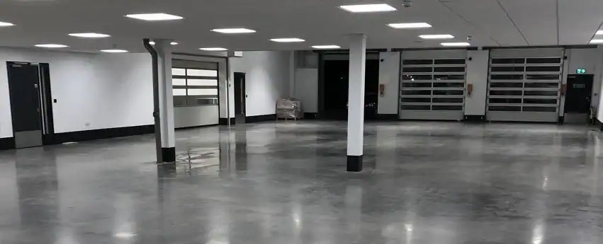 A large, empty industrial garage in Middlesex & New London County, CT, featuring polished concrete floors, white walls, ceiling lights, several support columns, multiple closed garage doors, and a small stack of items near the back wall.
