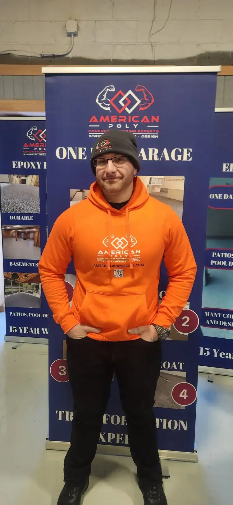 A person wearing an orange hoodie and black pants stands smiling in front of a blue American Poly banner with information about epoxy flooring Hartford and garage transformations, serving Middlesex & New London County and New Haven.