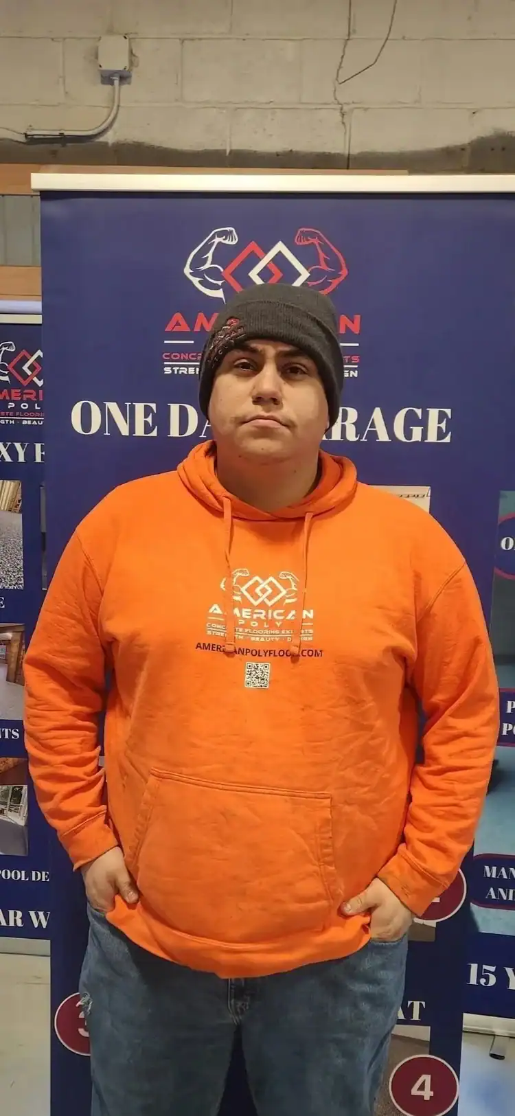 A person in an orange hoodie and gray beanie stands before a blue "One Day Garage" banner, featuring American Polyfloor logos and info on epoxy flooring Hartford, CT—serving Middlesex & New London County.