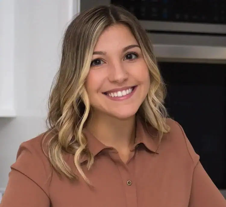 A woman with wavy blonde hair, wearing a brown button-up shirt, smiles warmly at the camera while standing indoors in front of a kitchen oven in Middlesex & New London County, CT.