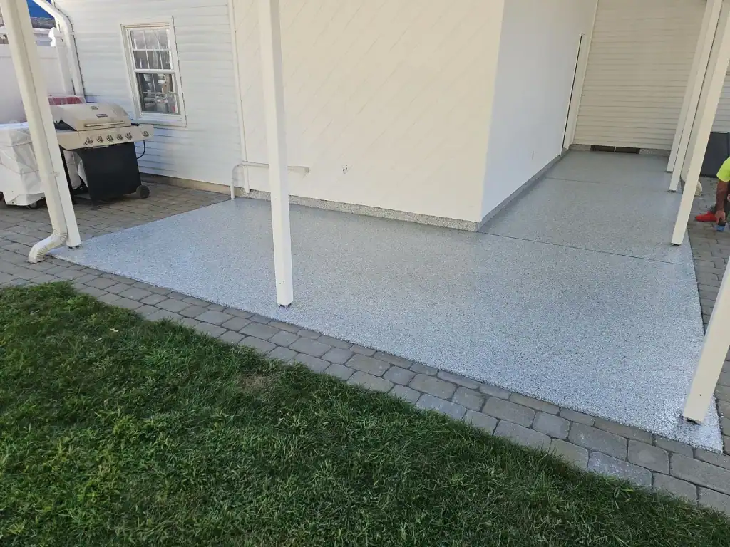 A clean, light gray patio with a speckled finish&mdash;reminiscent of epoxy flooring Hartford or New Haven, CT residents love&mdash;sits next to a building with white siding. The patio is bordered by a stone walkway and green grass, with a grill visible on the left.