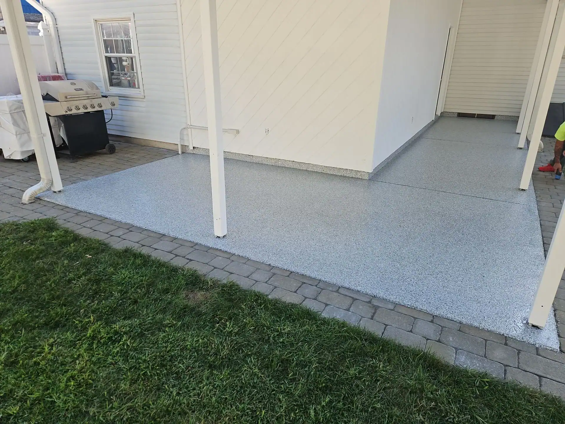 A clean, light gray patio with a speckled finish—reminiscent of epoxy flooring Hartford or New Haven, CT residents love—sits next to a building with white siding. The patio is bordered by a stone walkway and green grass, with a grill visible on the left.