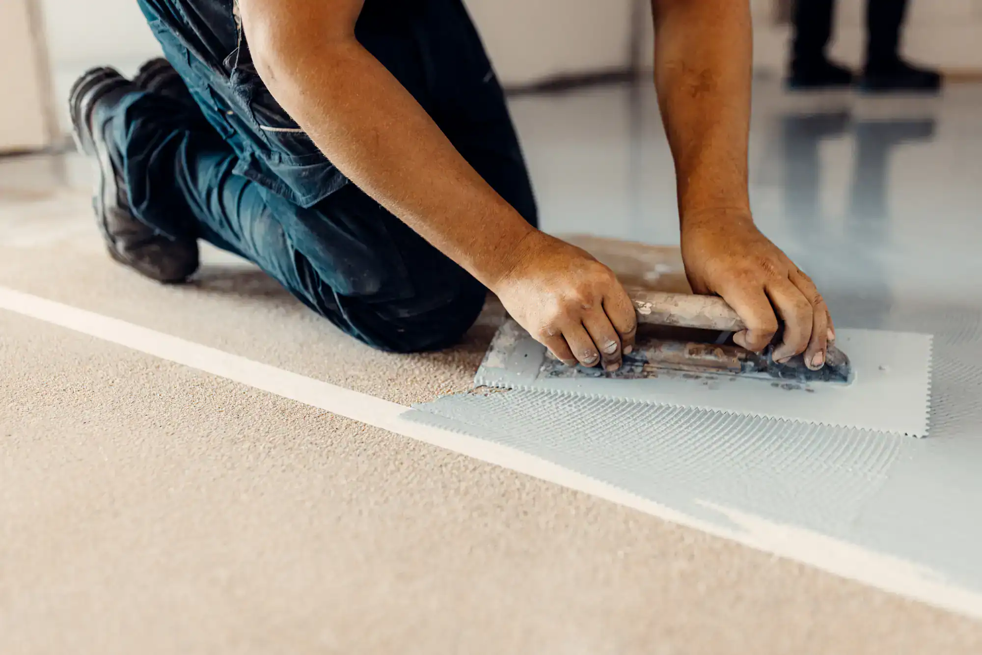 A person kneels on the floor, using a trowel to spread adhesive or leveling compound on a surface, preparing it for flooring installation. The worker wears dark pants and the focus is on their hands and tool.