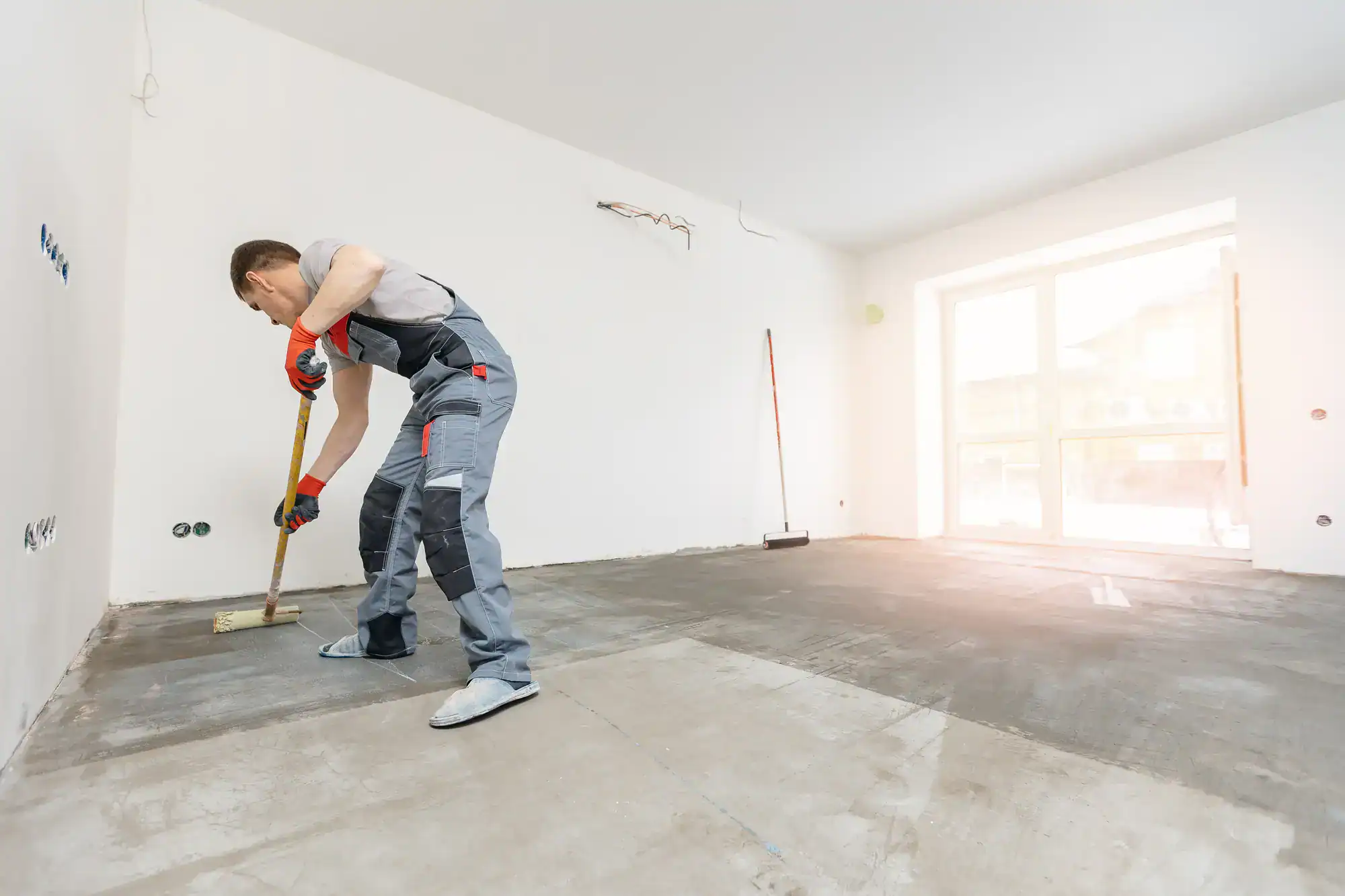 A person in work overalls uses a broom to sweep the floor of an unfinished, empty room with white walls and large windows, preparing the space for renovation.