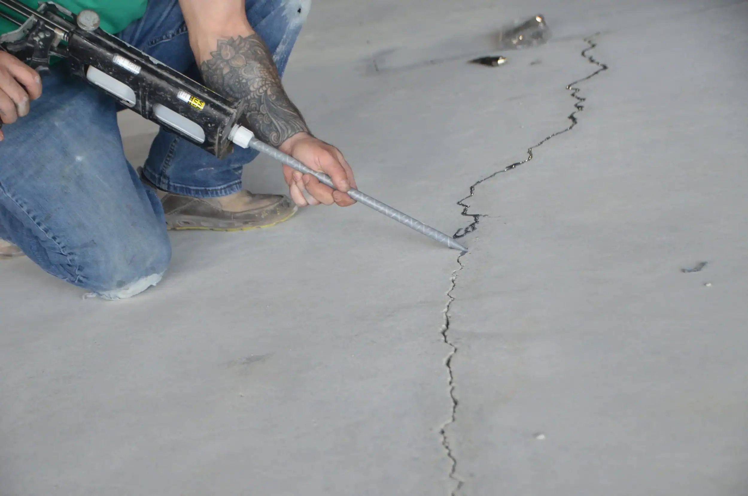 A person in jeans and work boots kneels on a concrete floor in East Hartford, CT, using a caulking gun to fill a long crack—showcasing skilled prep work for professional epoxy flooring. Their tattooed arm is visible as they work.