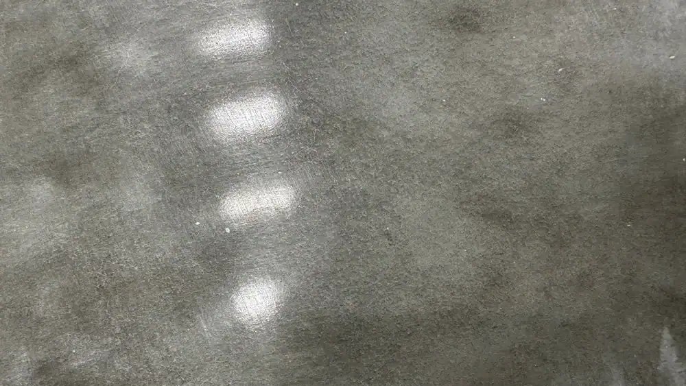 A close-up of a smooth concrete surface with a few scattered white patches and reflections, resembling the mottled, shiny look often seen in epoxy flooring Hartford or Middlesex & New London County installations.