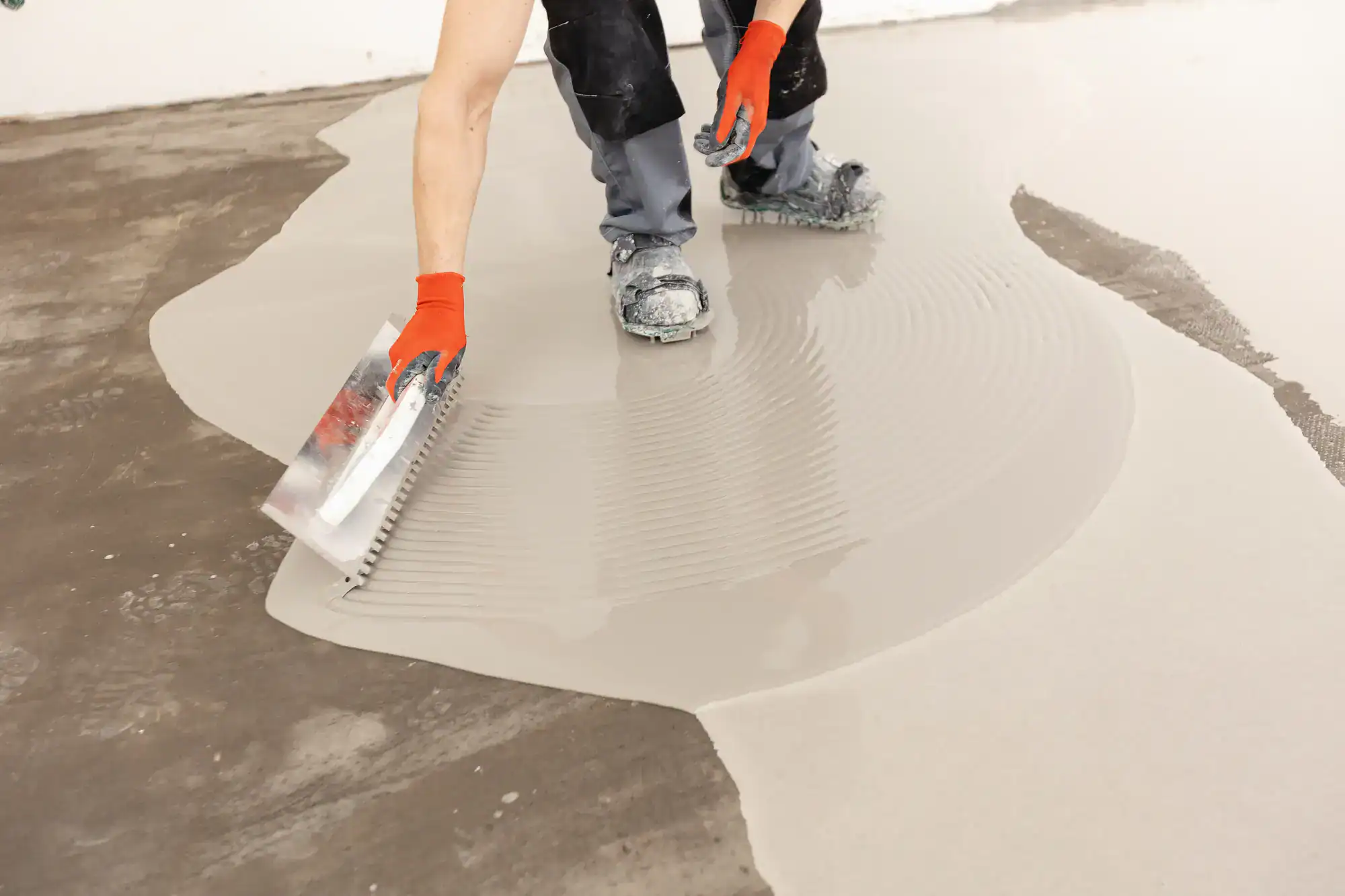 A person wearing orange gloves and work pants uses a notched trowel to spread self-leveling concrete over a floor, creating smooth, even lines on the surface.