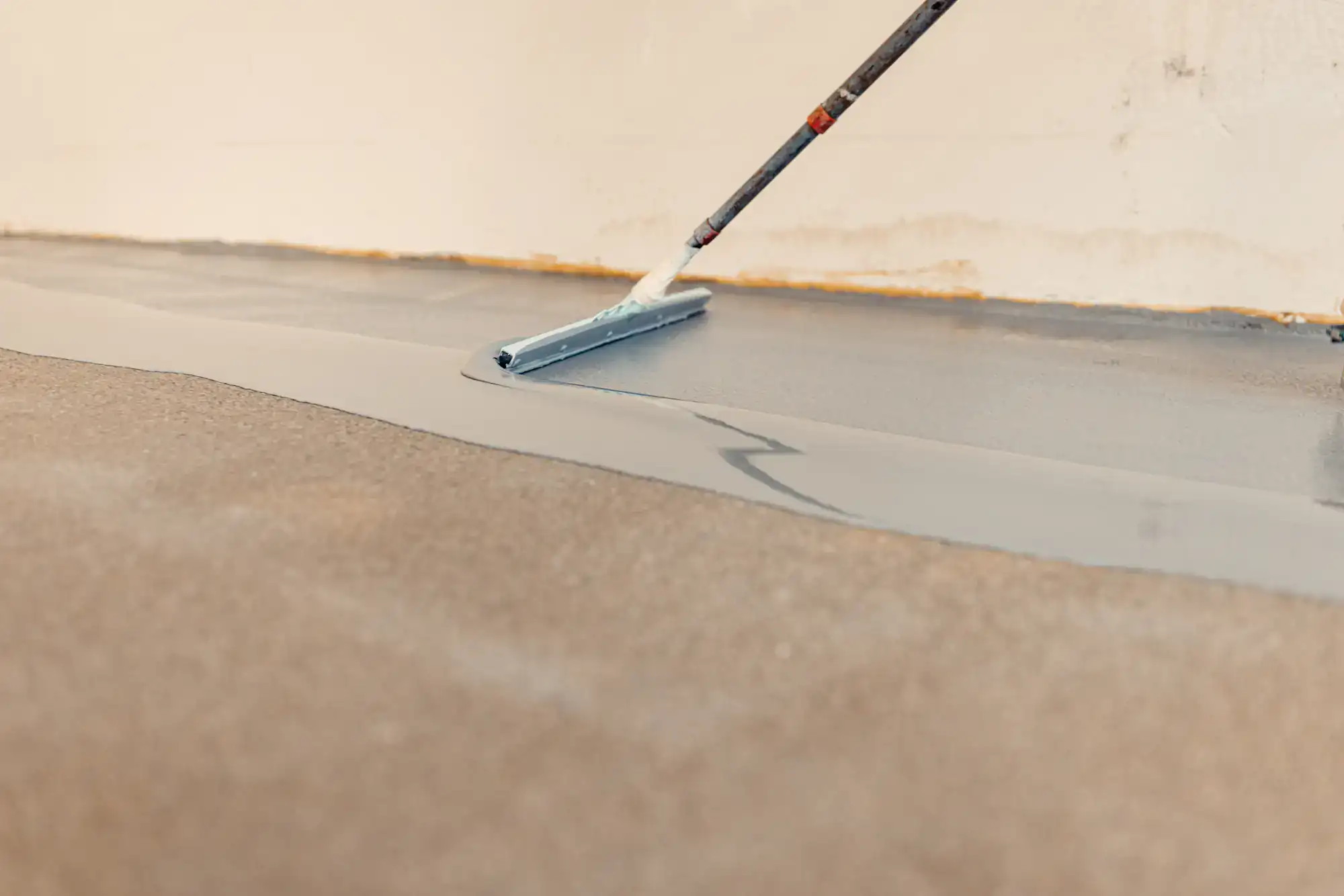 A close-up of a person using a long-handled tool to spread a layer of gray floor coating or paint over a rough concrete surface near a wall.