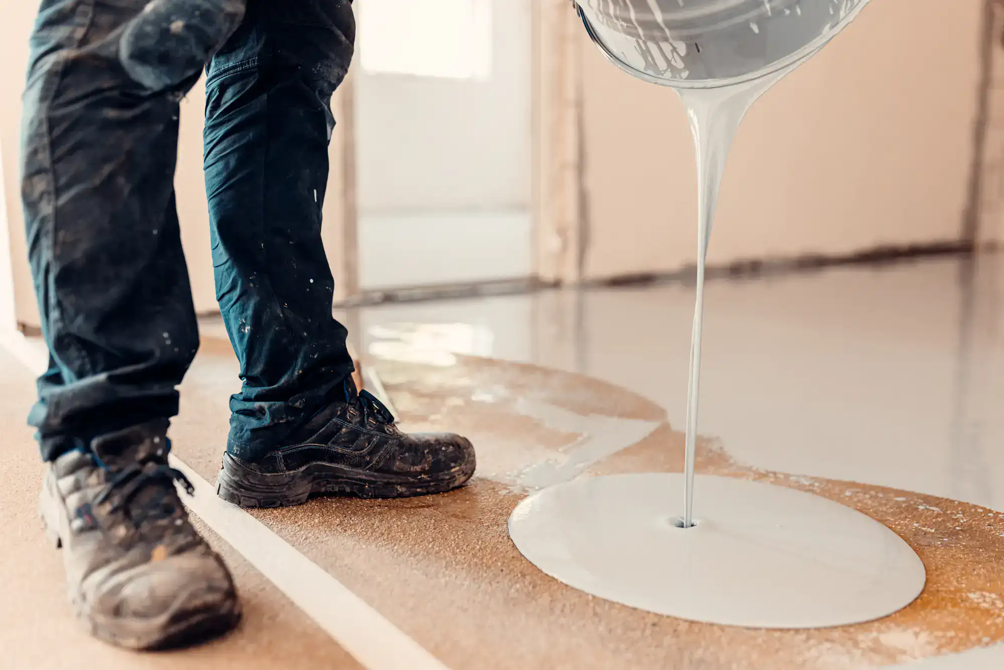 A person wearing work boots and dark pants pours liquid white floor leveling compound onto a floor inside a building under renovation.