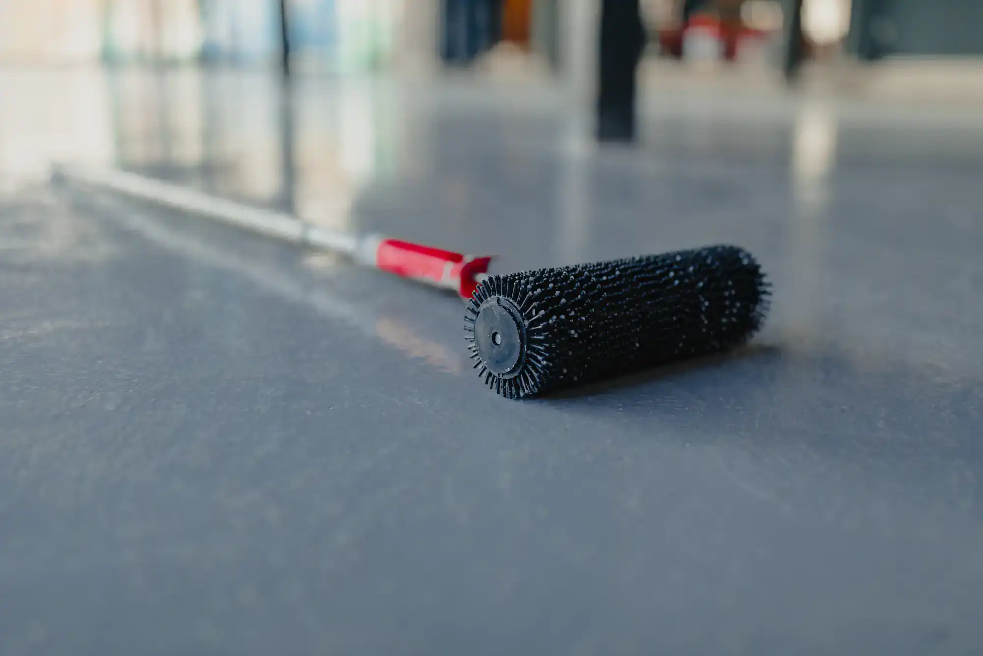 A close-up of a spiked roller tool with a red handle lying on a smooth, shiny gray floor, used for leveling or treating flooring surfaces.