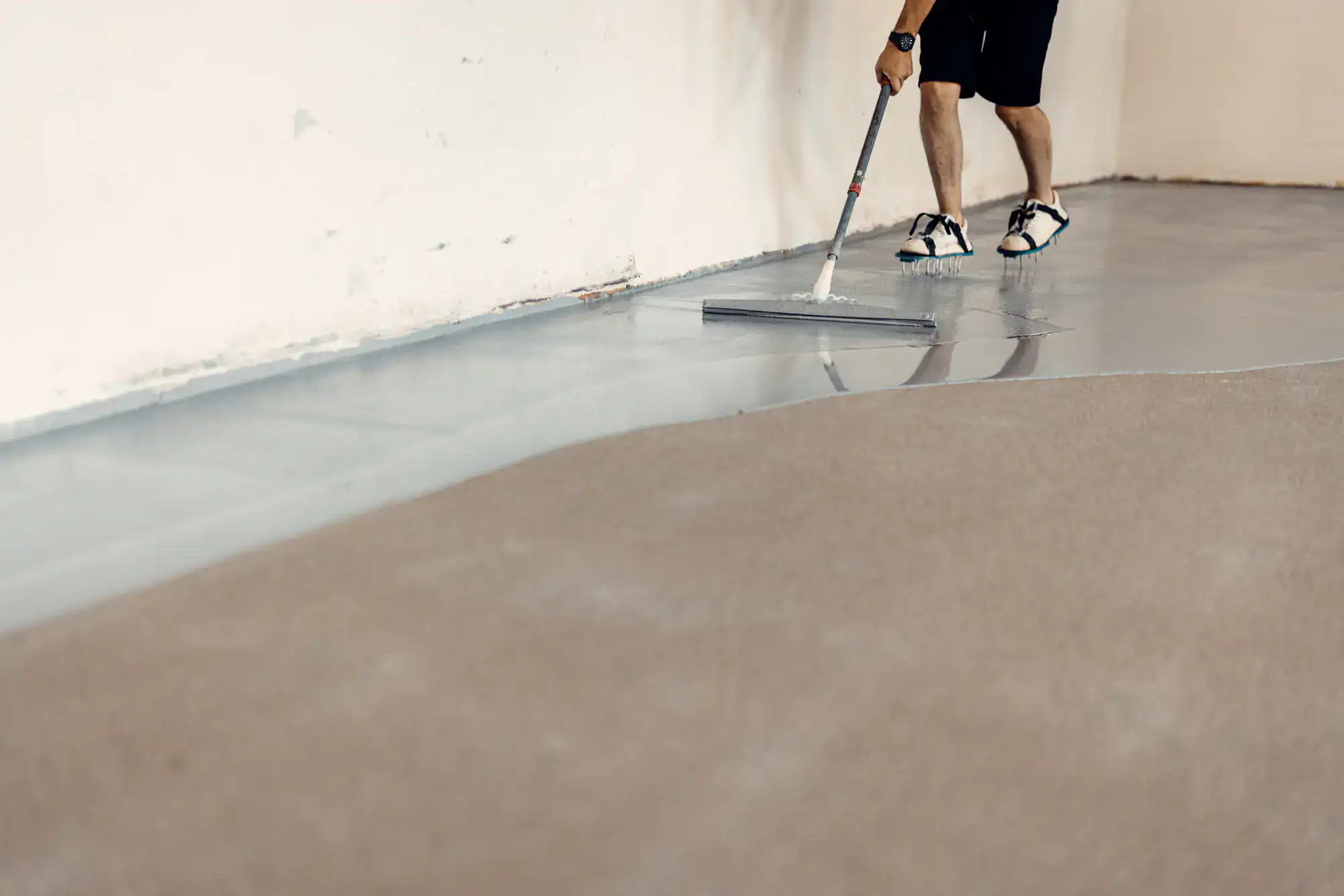 A person wearing sandals and shorts spreads a liquid coating on a floor with a long-handled tool, preparing the surface next to a wall and an unfinished section.