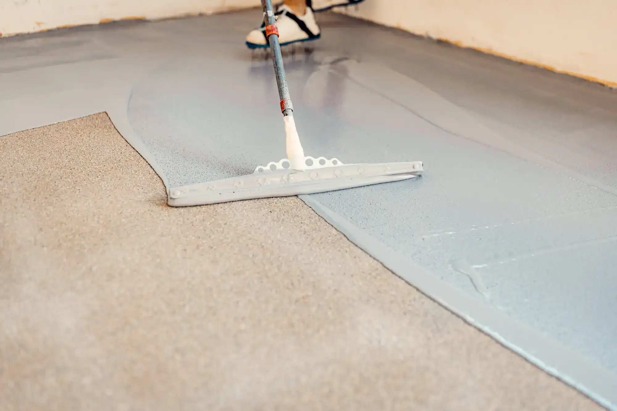 A person uses a large squeegee to spread gray epoxy floor coating over a concrete surface, partially covering the textured floor in a smooth, shiny layer.