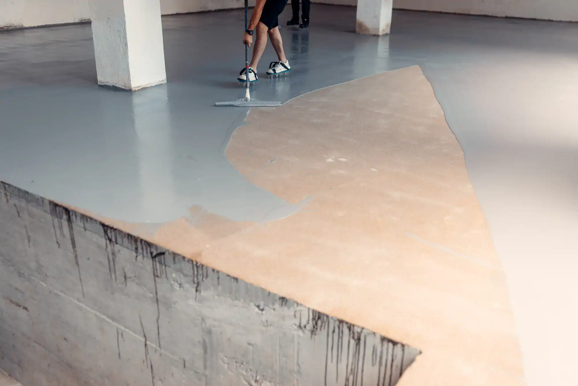 A person uses a tool to spread gray epoxy coating over a concrete floor in an unfinished indoor space with white columns. The coating covers about half of the visible floor.