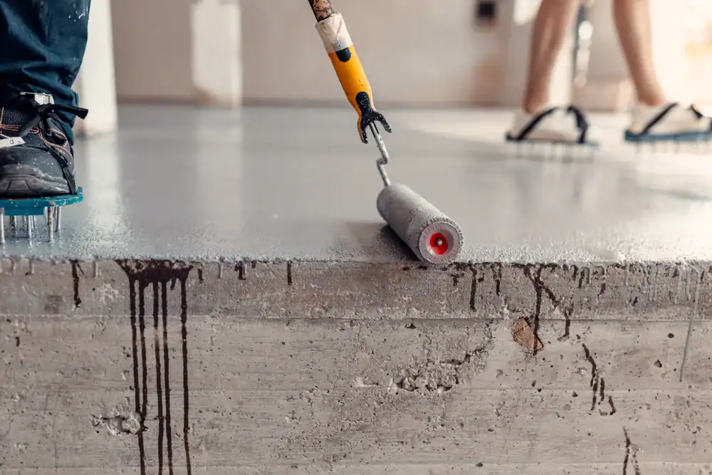 A close-up of a person applying epoxy flooring on a gray concrete floor in Hartford, New Haven, Middlesex & New London County, with another person’s legs visible in the background and dark paint drips on the concrete edge.