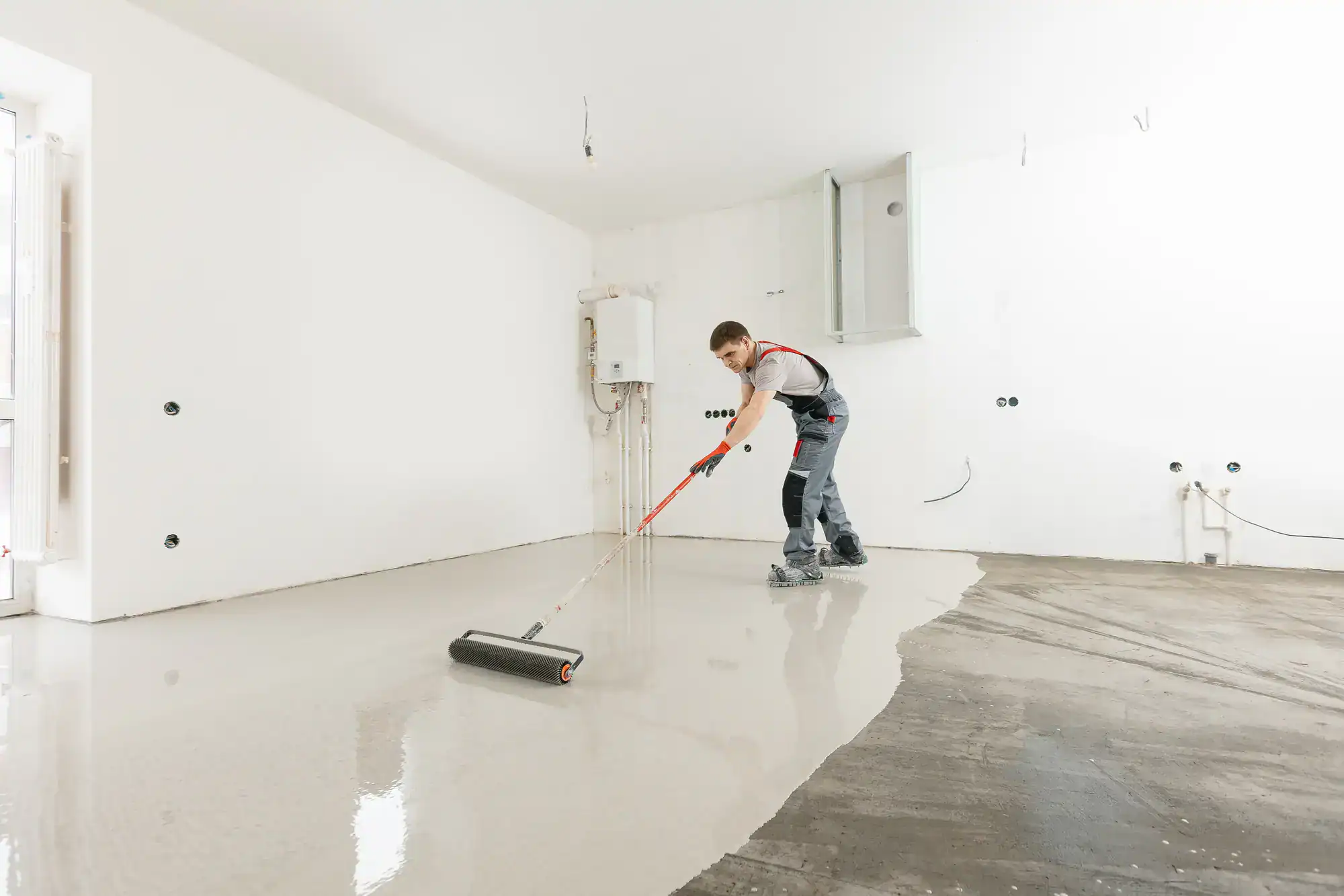 A worker in overalls spreads wet concrete on a floor using a long-handled roller in a bright, unfinished room with white walls and exposed pipes.