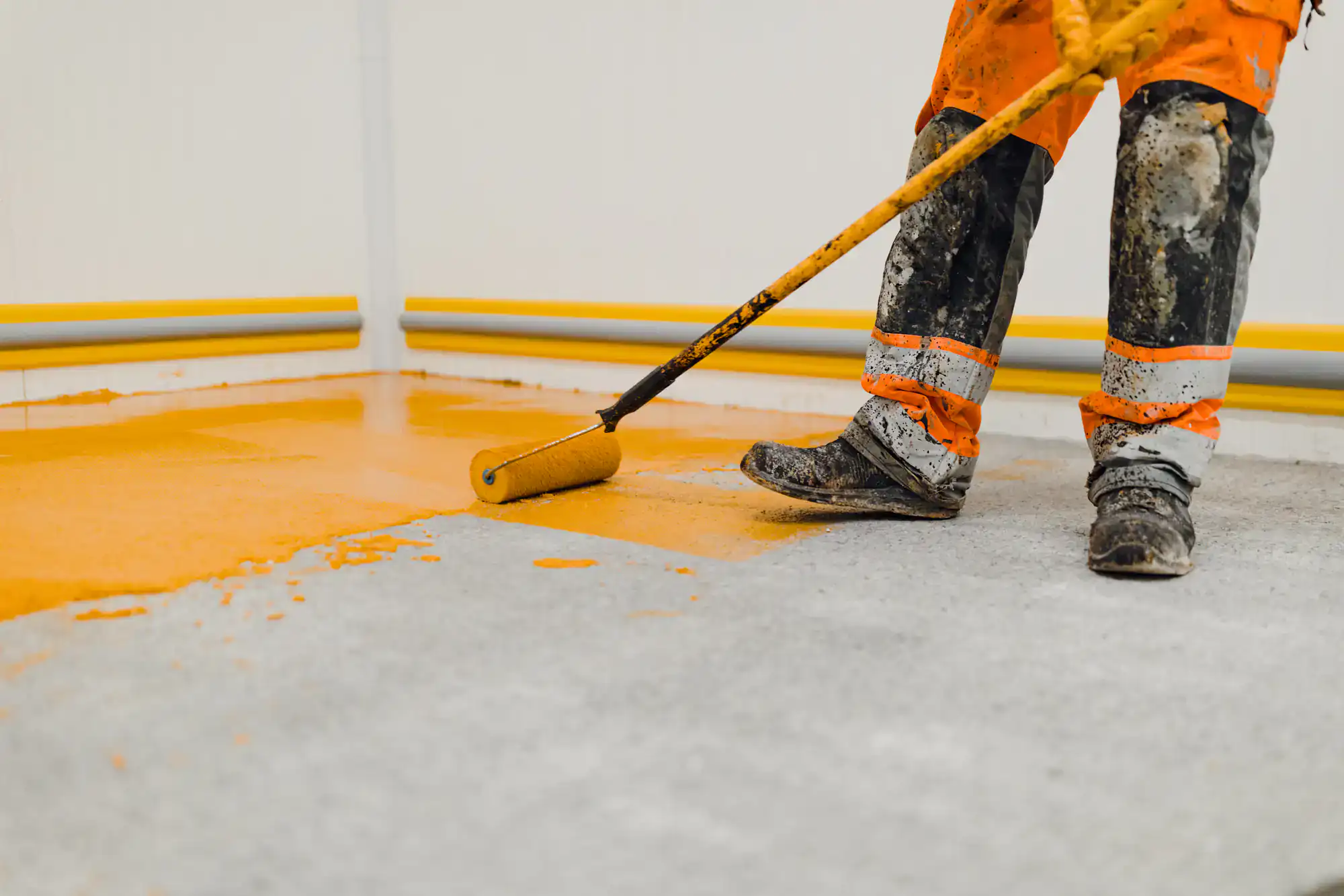 A worker in orange safety pants and muddy boots uses a paint roller to apply yellow paint to a concrete floor near a corner with yellow piping along the walls.