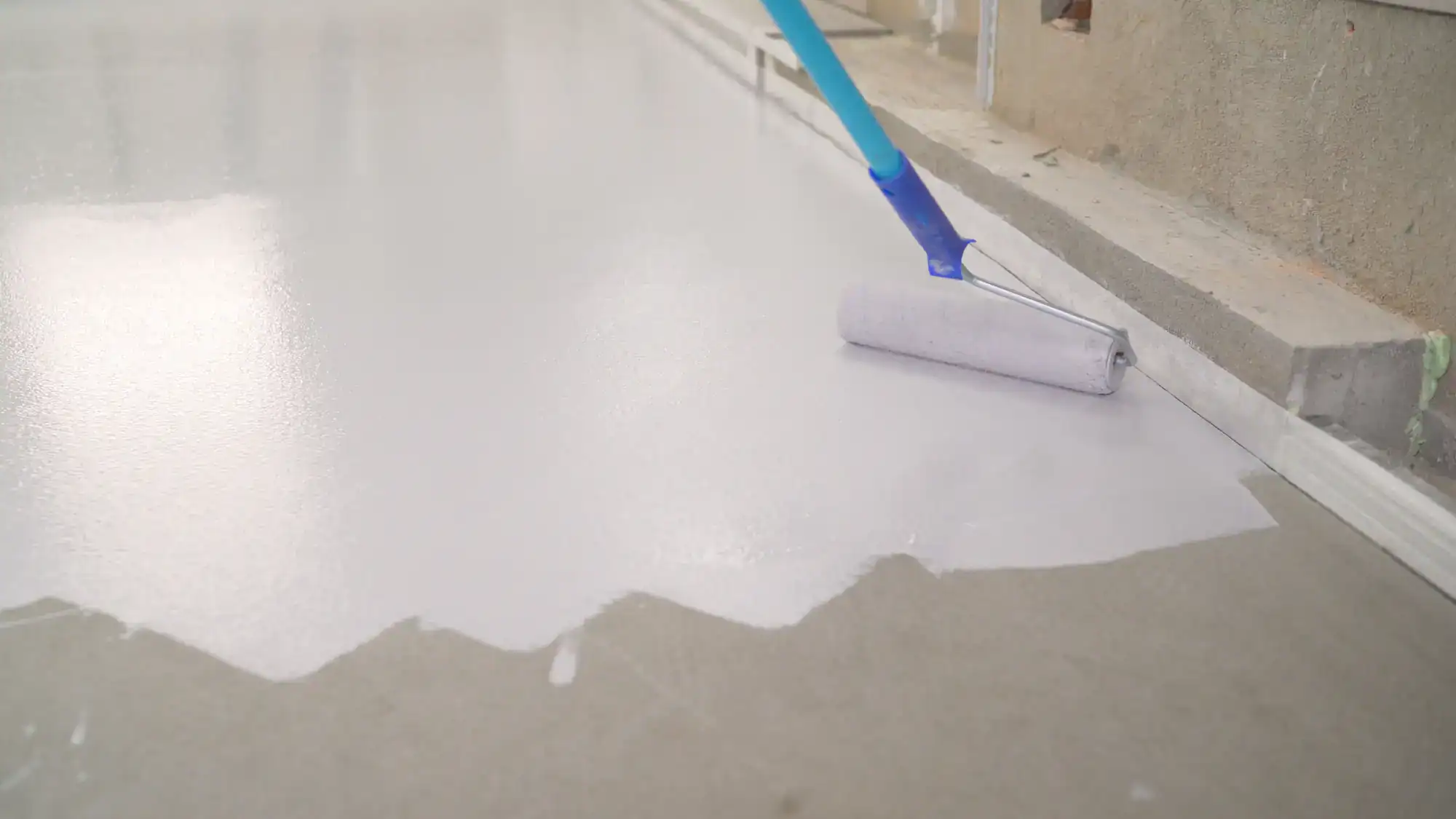 A close-up of a paint roller with a blue handle applying white paint or primer to a concrete floor in an indoor setting. The roller moves from unpainted to freshly coated surface.
