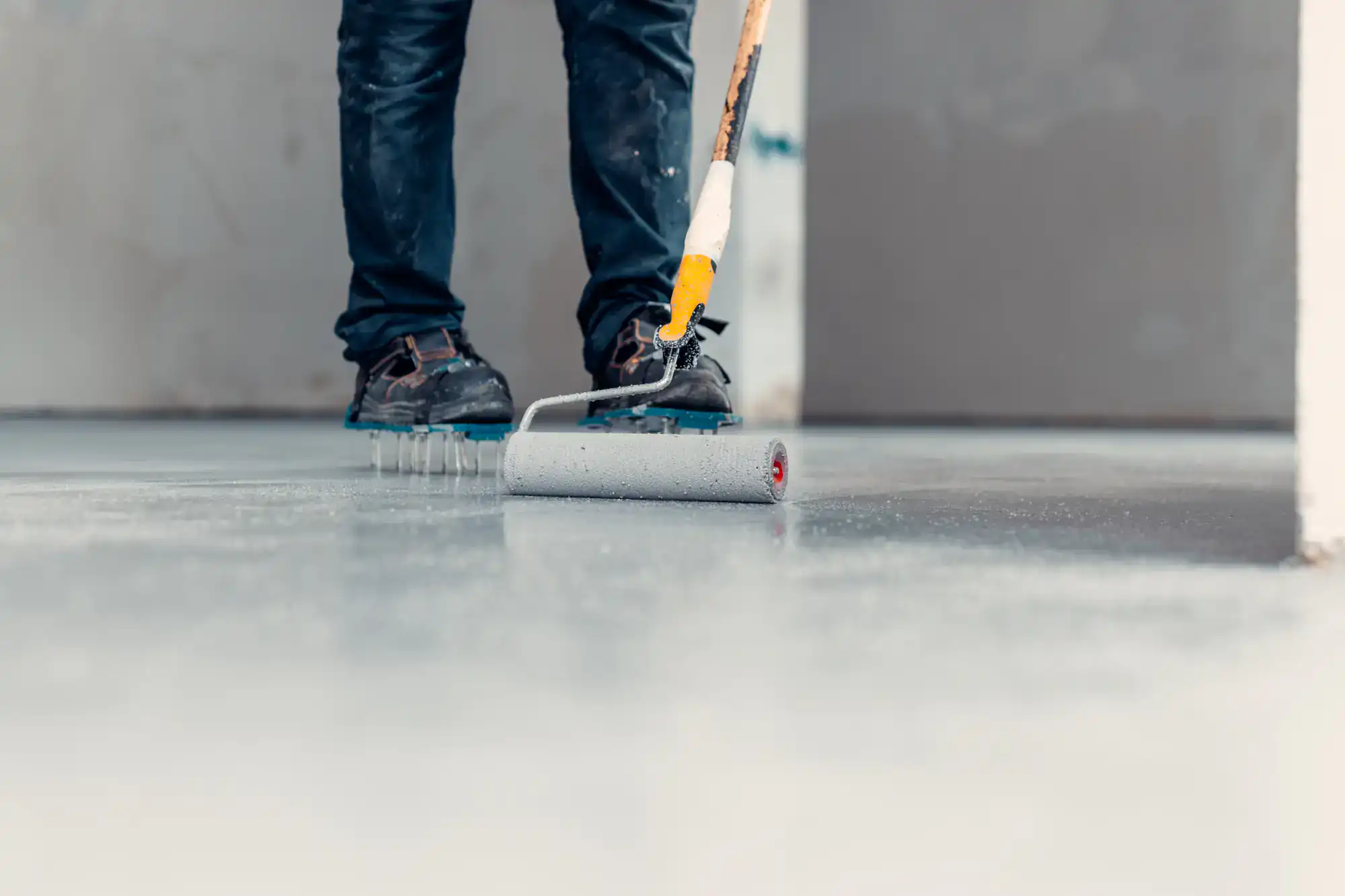 A person wearing dark pants and work shoes uses a roller brush with a long handle to paint or coat a concrete floor inside a building. The focus is on the roller and the lower part of the person's legs.
