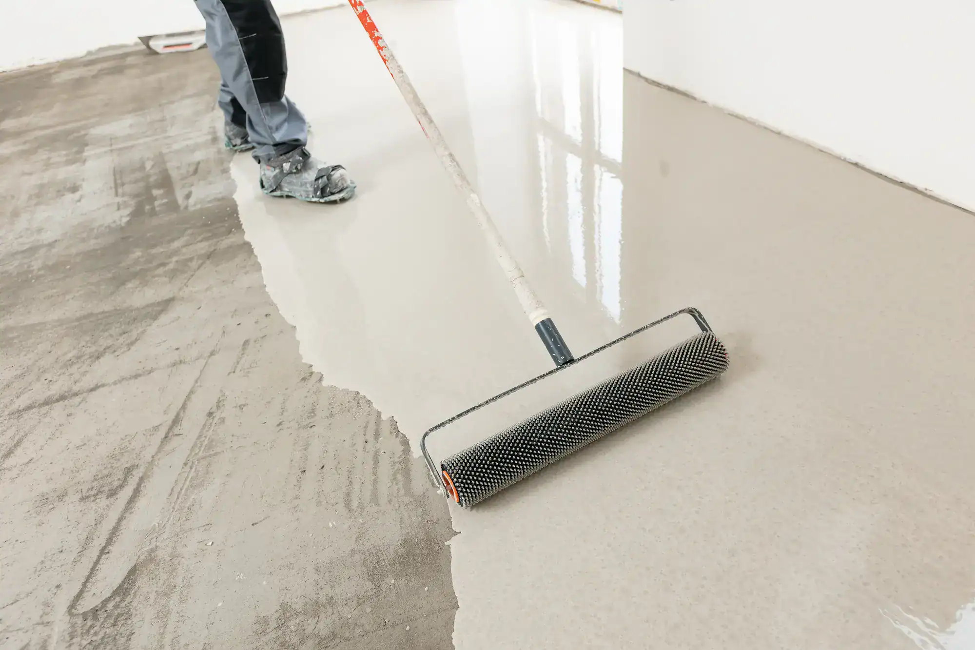A person uses a spiked roller to smooth and level freshly poured concrete or self-leveling compound on a floor, with a visible line between the wet and untreated surface.