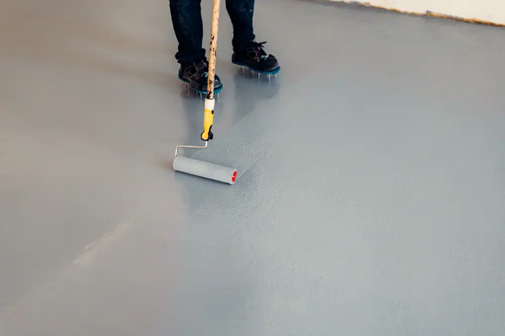 A person in Middlesex & New London County uses a paint roller with an extension handle to apply gray paint or coating to a floor. Only the lower legs and feet of the individual are visible.