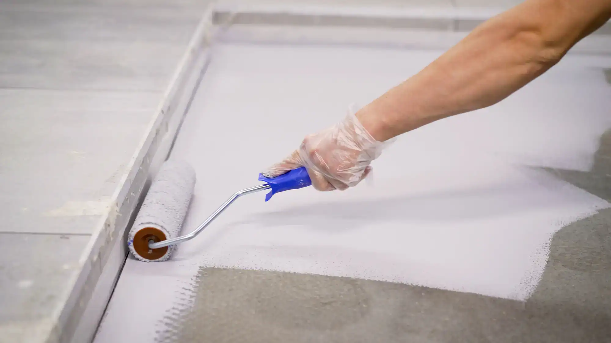 A person wearing a plastic glove uses a blue-handled paint roller to apply white paint or primer to a concrete floor.