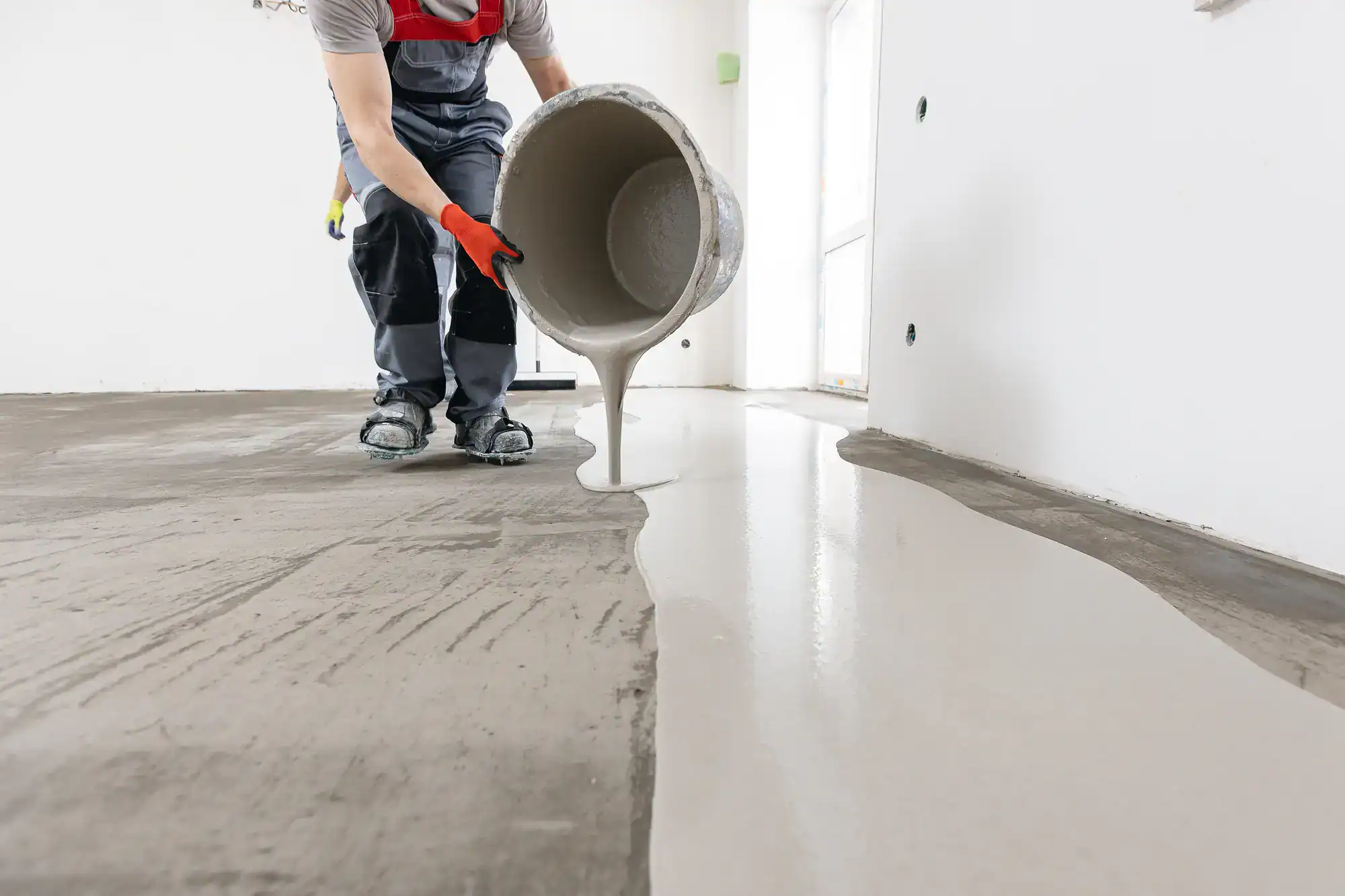 A worker in protective gear pours liquid cement or self-leveling compound onto a floor in a bright, unfinished room during a flooring installation or renovation.
