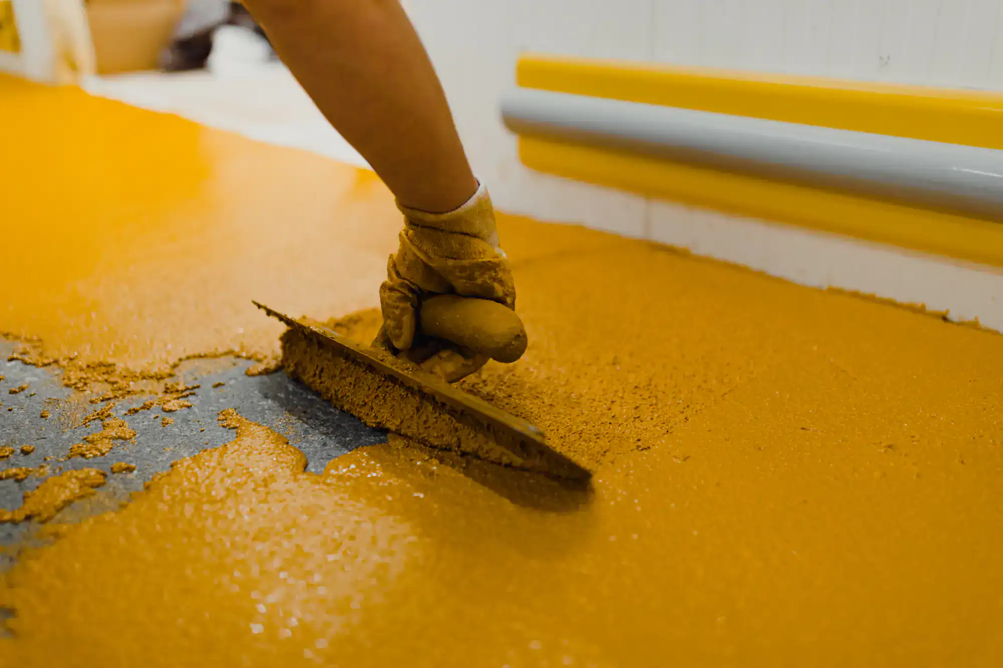 A gloved hand uses a trowel to spread a layer of yellow epoxy flooring material over a surface, with a wall and baseboard visible in the background.