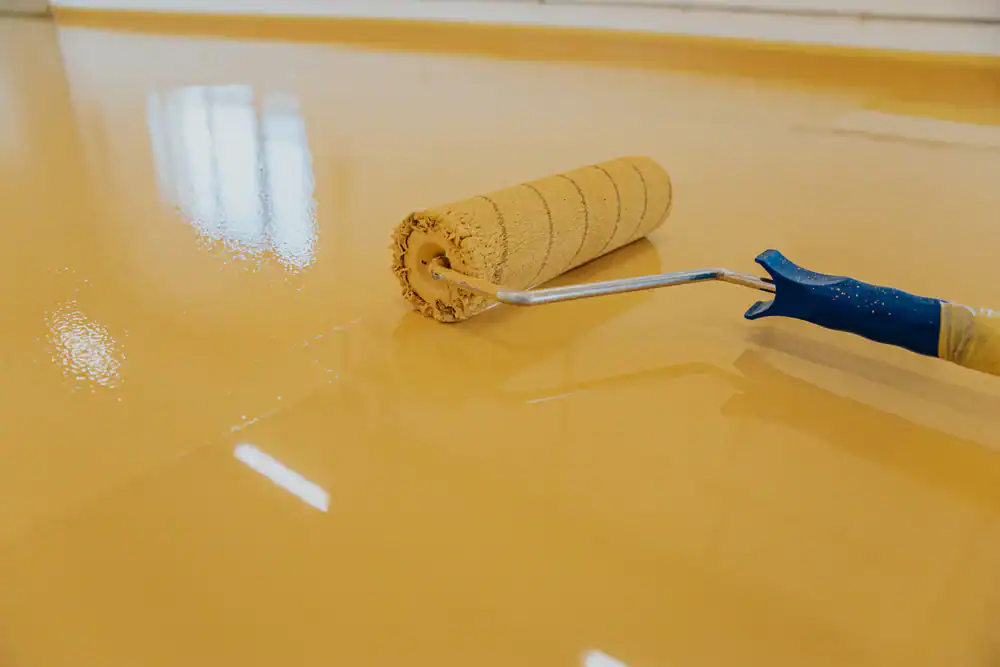 A paint roller with a blue handle is applying a smooth, glossy coat of yellow epoxy flooring in Hartford. Reflections of windows can be seen on the freshly painted, shiny surface&mdash;perfect for homes in Middlesex & New London County, CT.