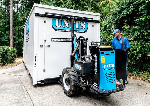 A worker in blue operates a small forklift labeled "UNITS" to move a portable storage container on a driveway surrounded by trees and greenery.