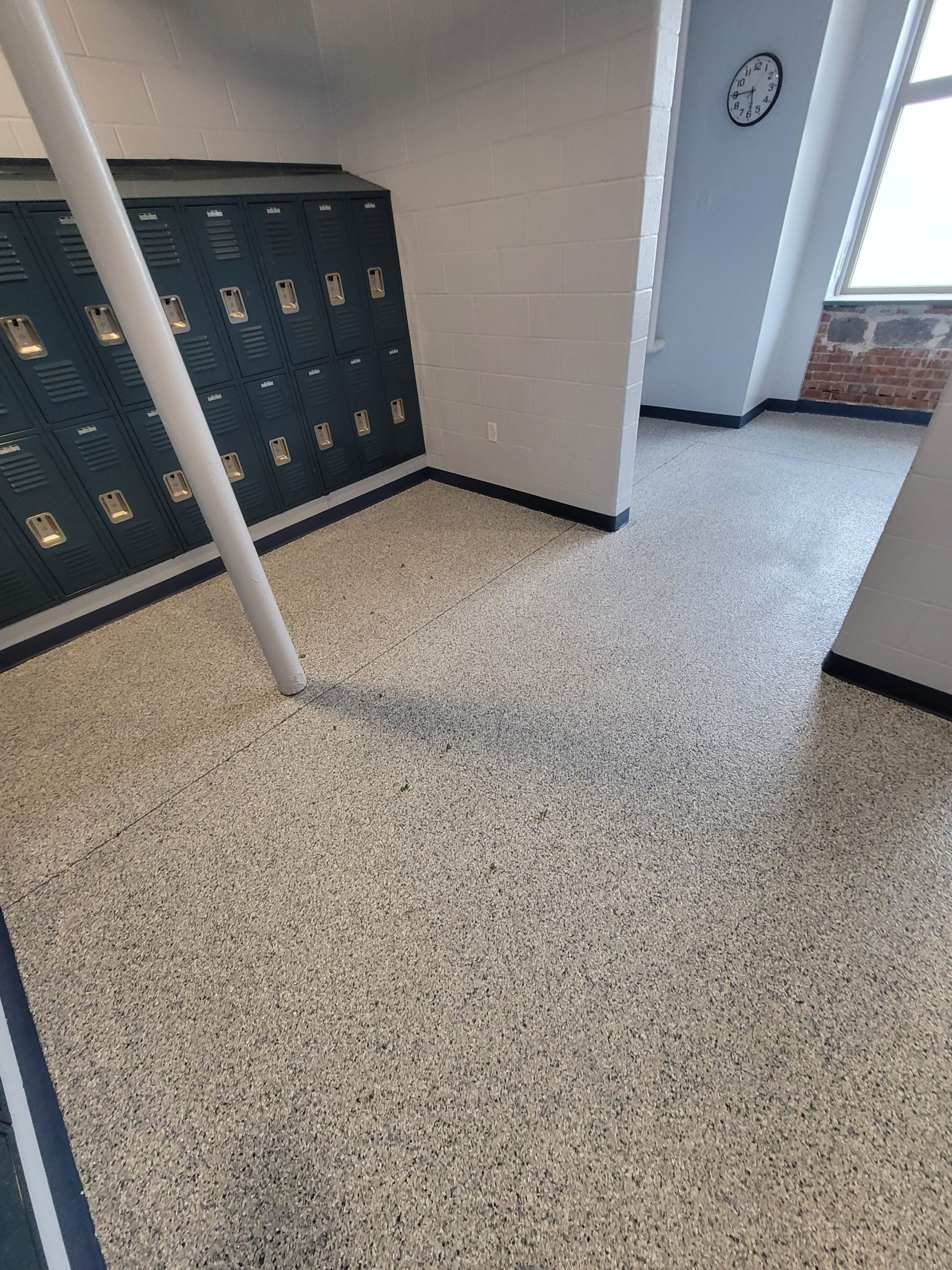 School hallway with a row of dark lockers, white pole, wall clock, window, and speckled flooring.