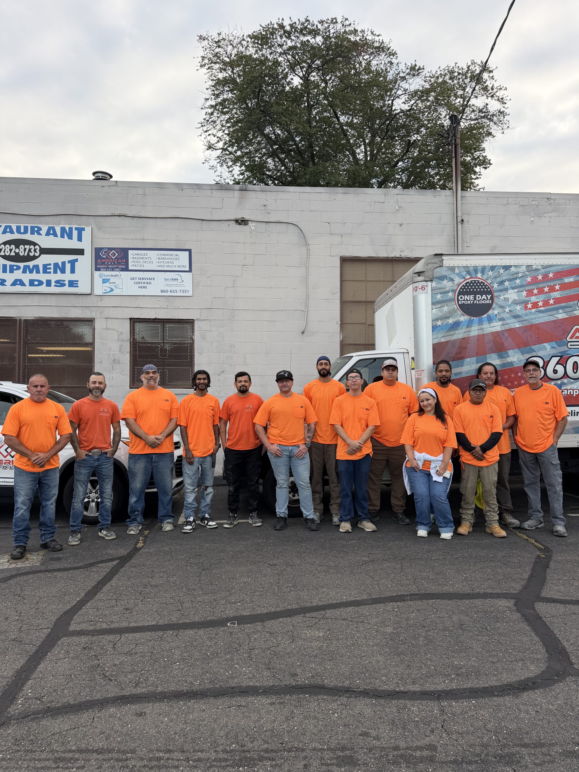 Fifteen people in orange shirts stand in a line outdoors by a white truck and building near a sign.