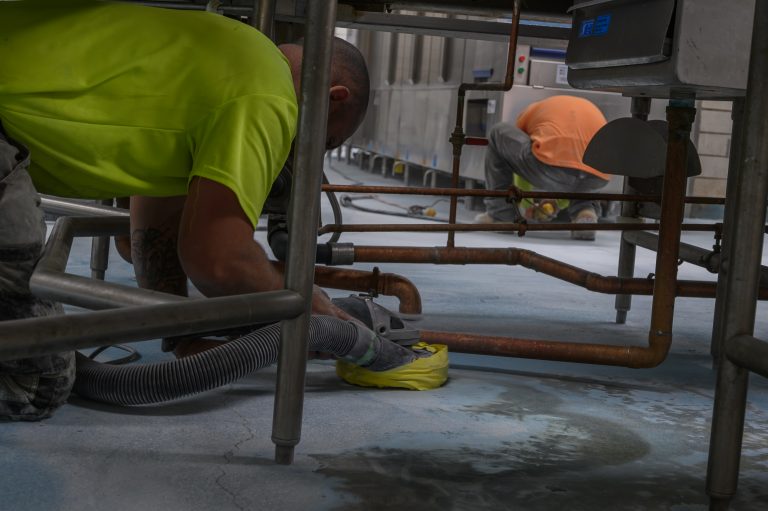 A worker in a neon shirt uses a power tool on copper pipes while another person works nearby.