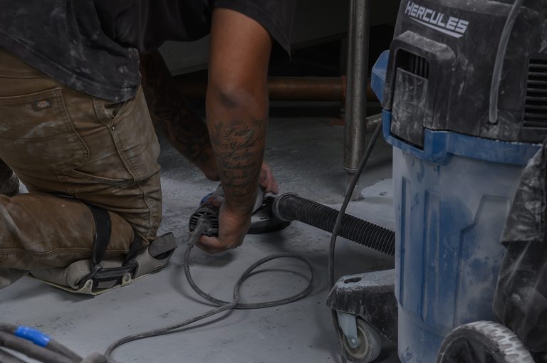 A worker kneels on a concrete floor using a handheld power sander connected to a vacuum.