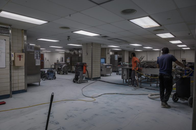Two workers clean and maintain equipment in a large commercial kitchen.