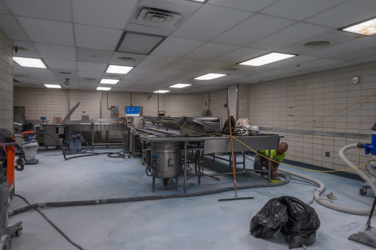 A worker uses a vacuum hose to clean the floor of a commercial kitchen.