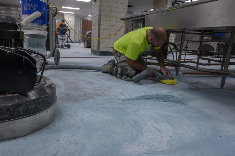 A worker in a neon yellow shirt uses a hand sander on a concrete floor in a commercial kitchen.