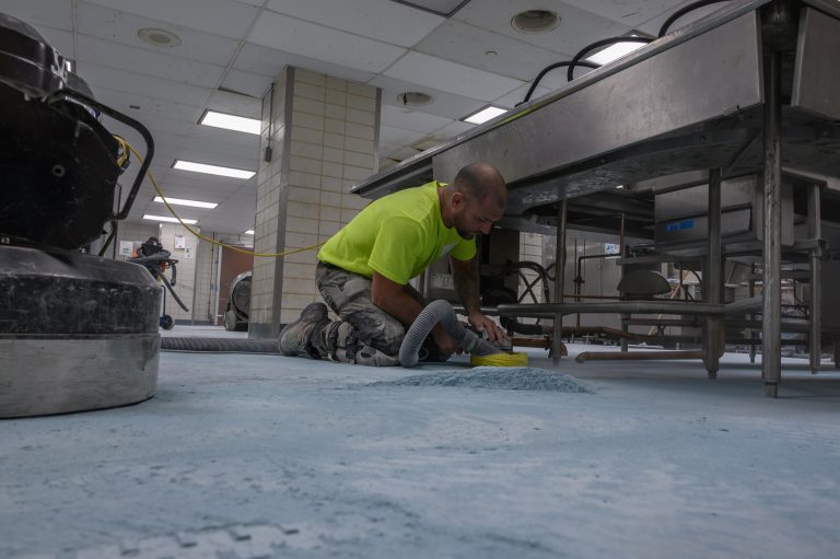 A construction worker kneels on a concrete floor using an industrial sander to smooth the surface.
