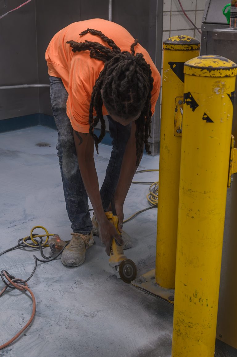 A worker in an orange shirt uses an angle grinder to cut the base of a yellow bollard.