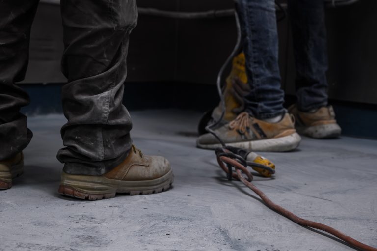Two construction workers wearing work boots stand on a concrete floor near an orange power cord.