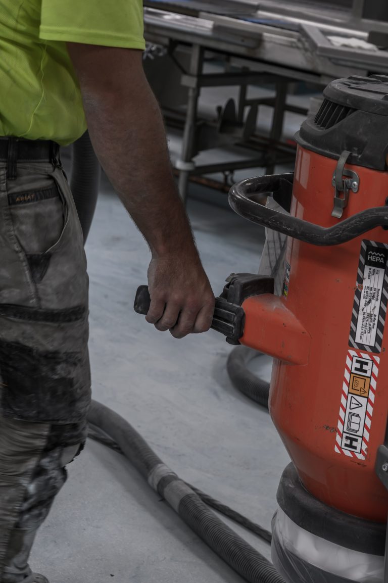 A worker in a yellow shirt pulls a large orange industrial HEPA vacuum across a concrete floor.