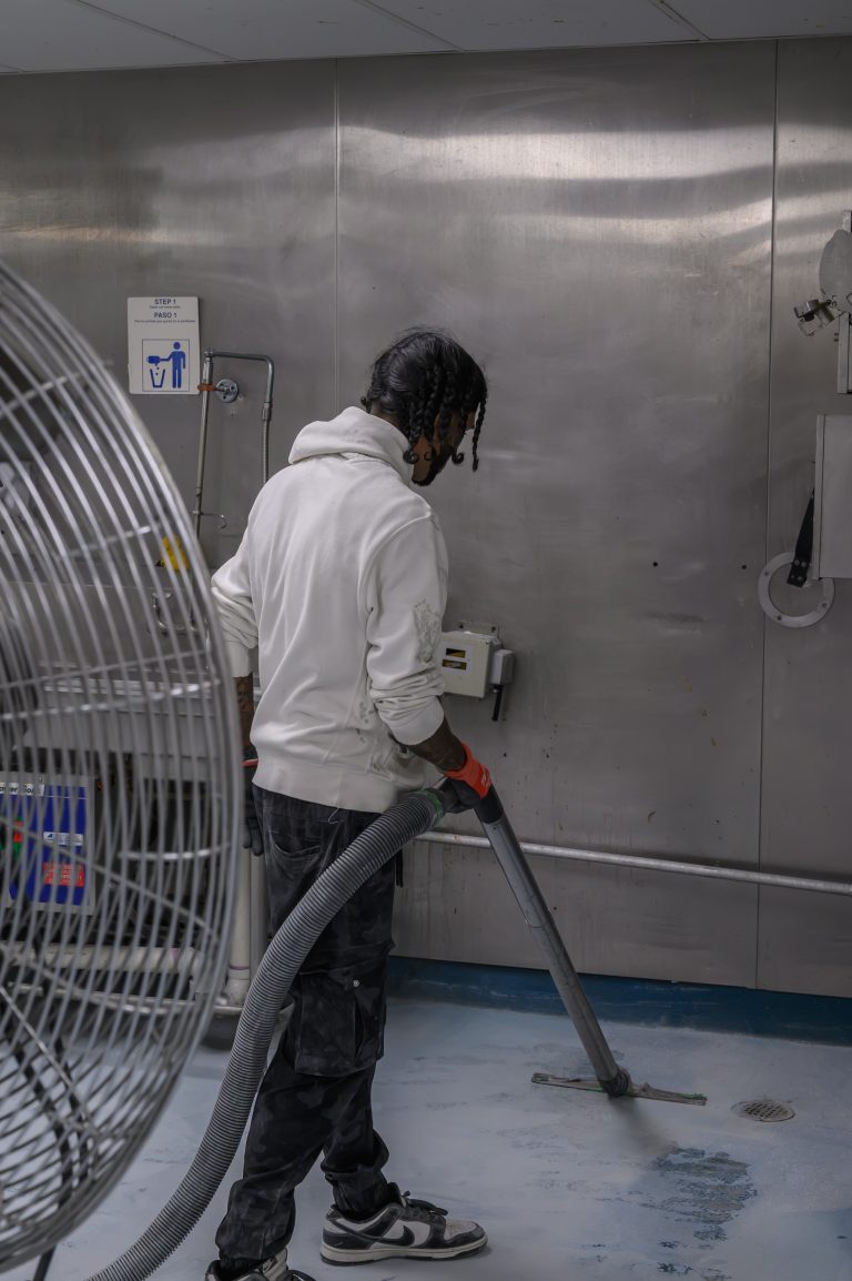 A person in a white hoodie vacuums the floor near metal walls and a large industrial fan.
